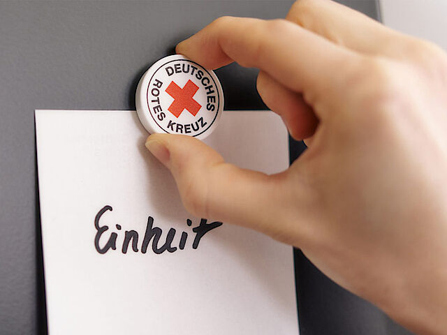 Jemand heftet mit einem Magnet mit DRKLogo ein Zettel mit dem Wort Einheit an die Wand